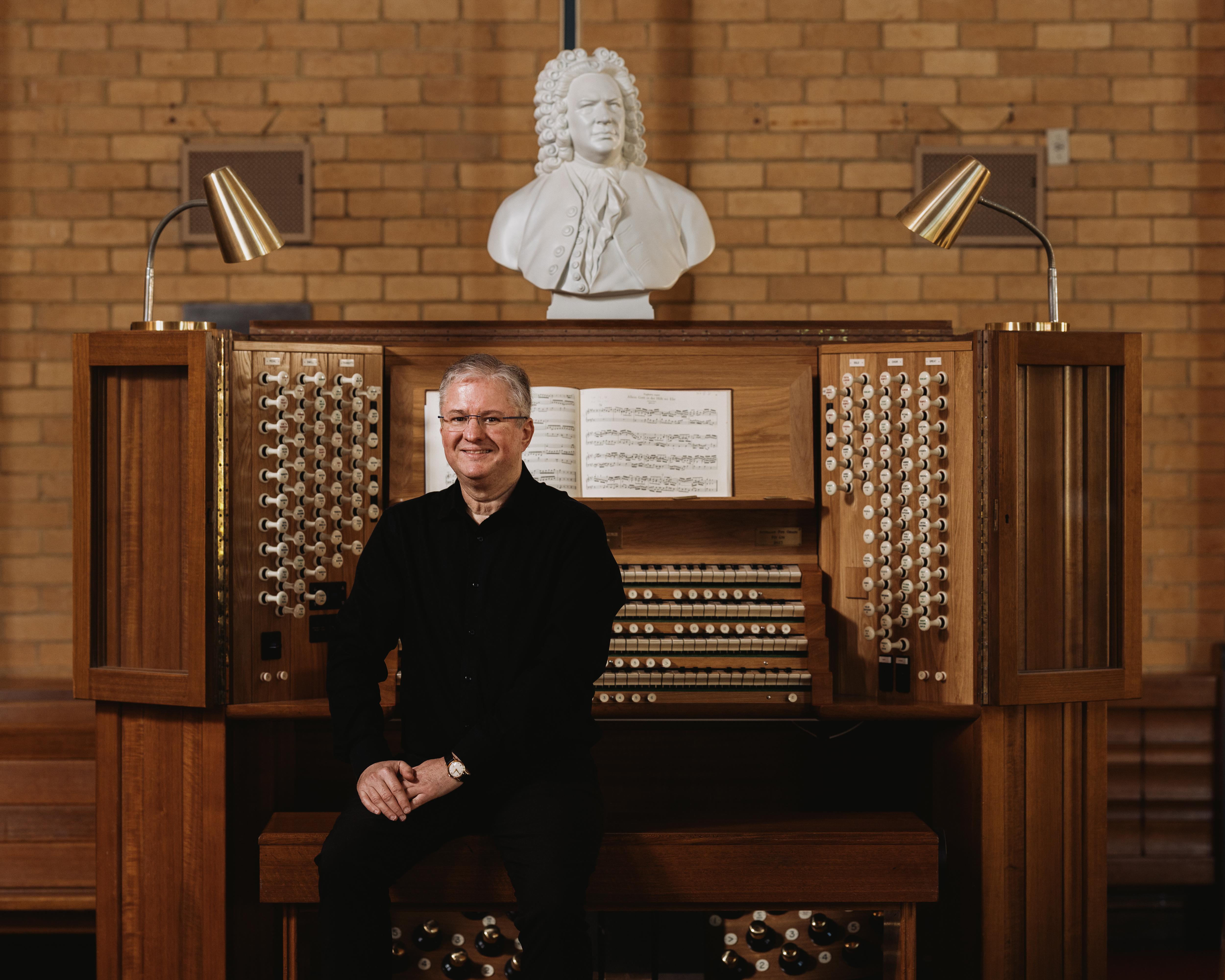 Organist seated at a large wooden pipe organ beneath a white bust and brick wall.