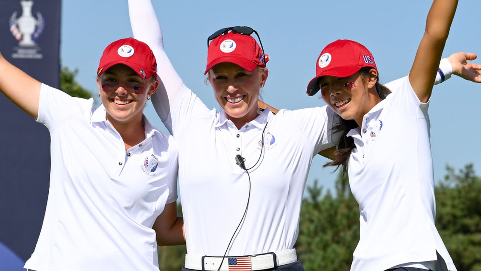 Three female golfers in white polos and red caps celebrate with their arms raised.