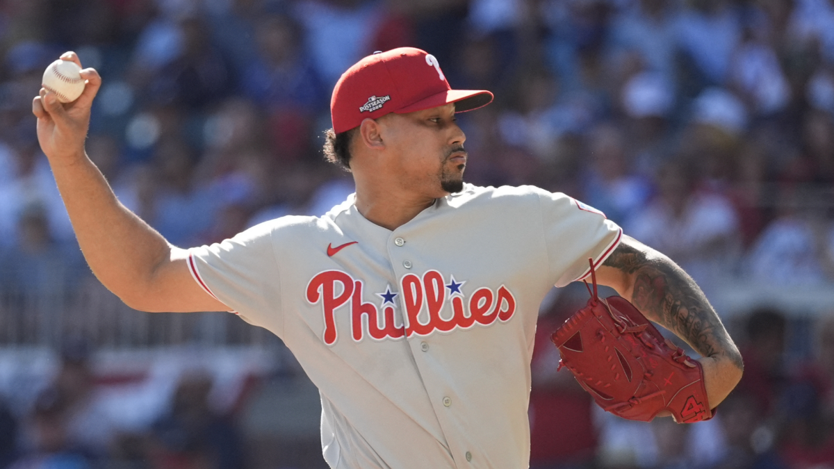 Phillies pitcher in a gray uniform raises a baseball, holding a red glove as he prepares to throw.