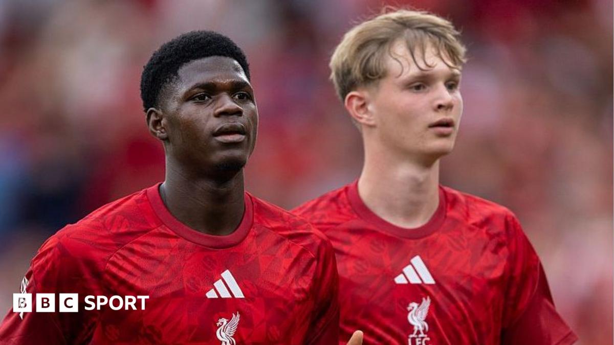 Two young footballers in red Liverpool shirts stand on a pitch against a blurred stadium crowd.