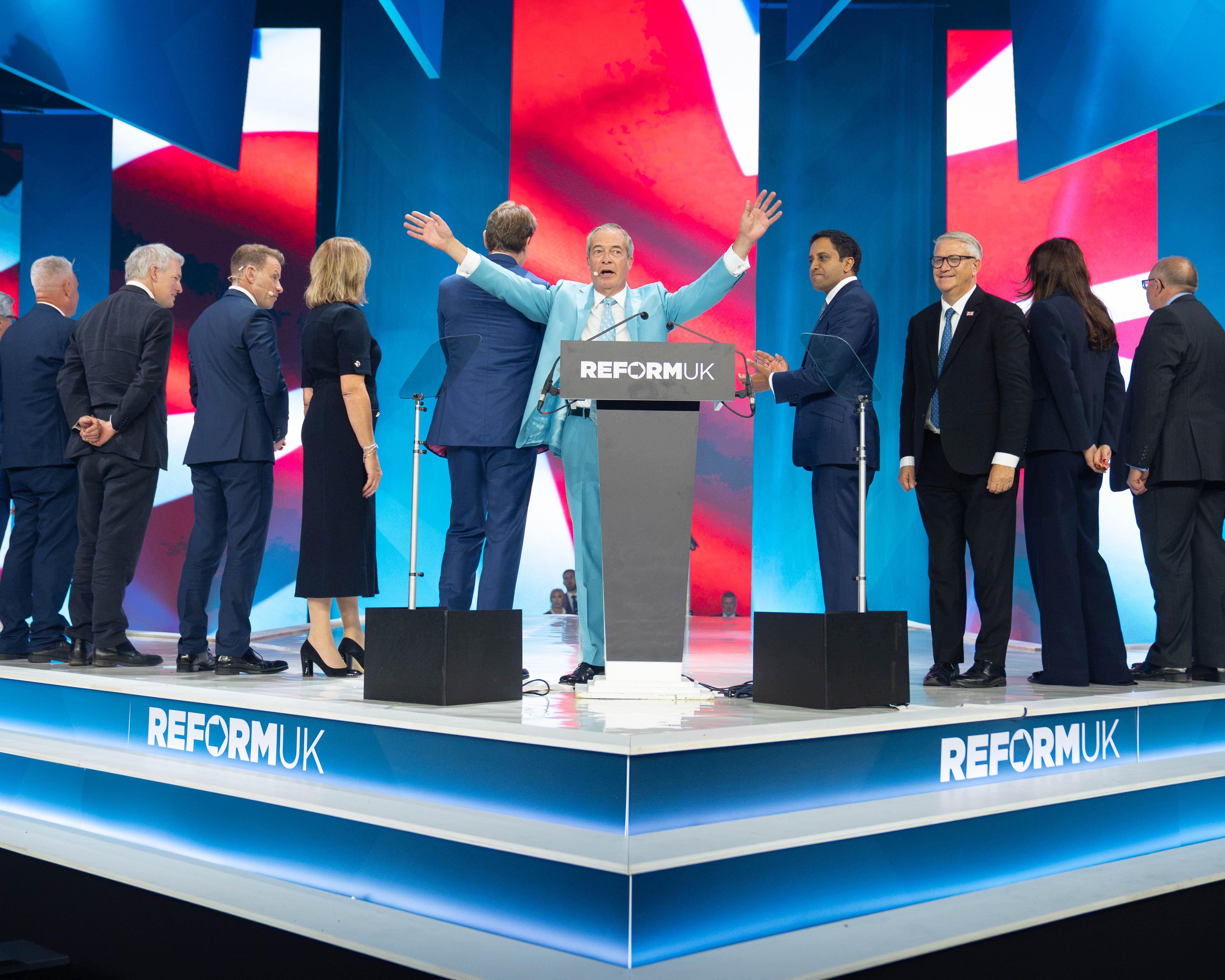 Reform UK leader speaks at a podium as party members stand onstage behind him.