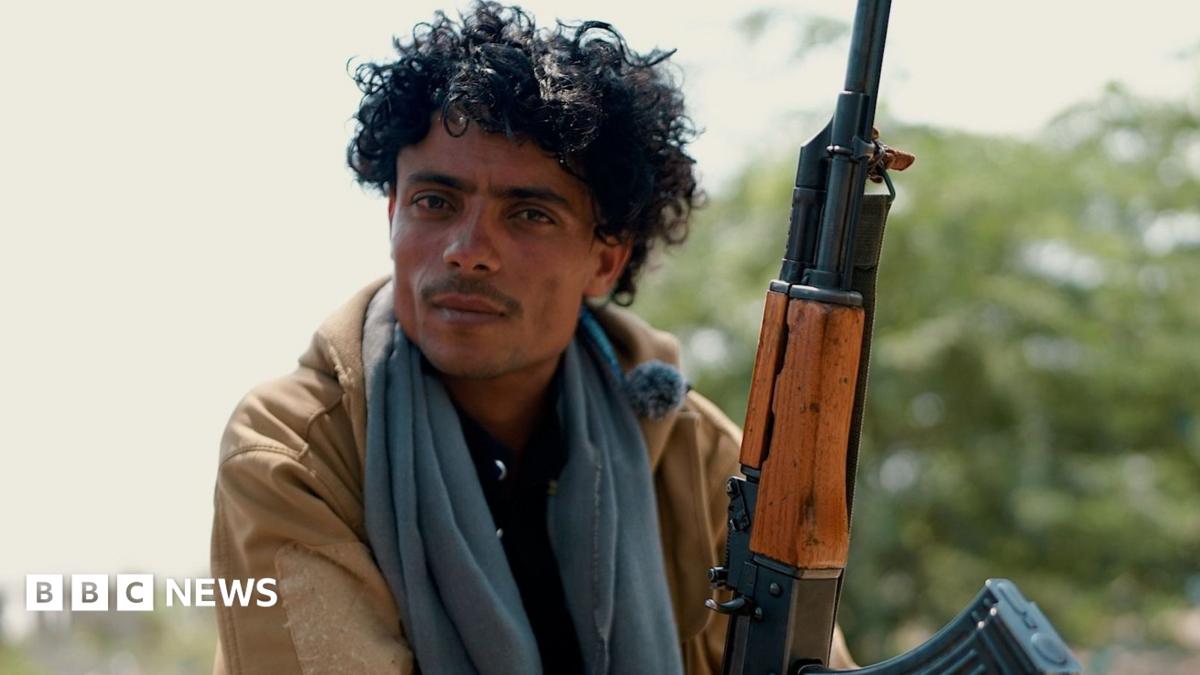 A curly-haired man in a tan jacket and gray scarf sits beside a rifle outdoors.