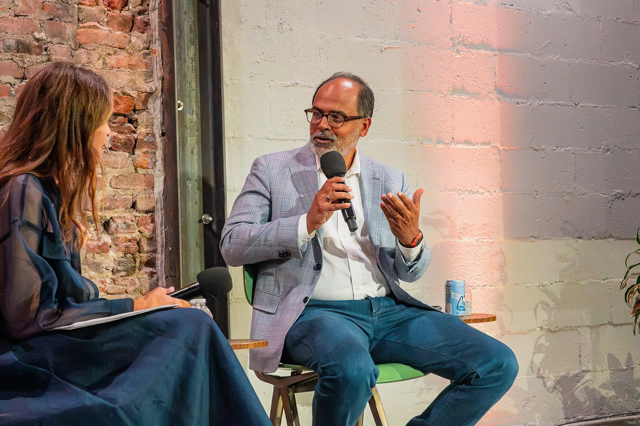 Man speaking into a microphone while a woman interviewer listens in an industrial interior.