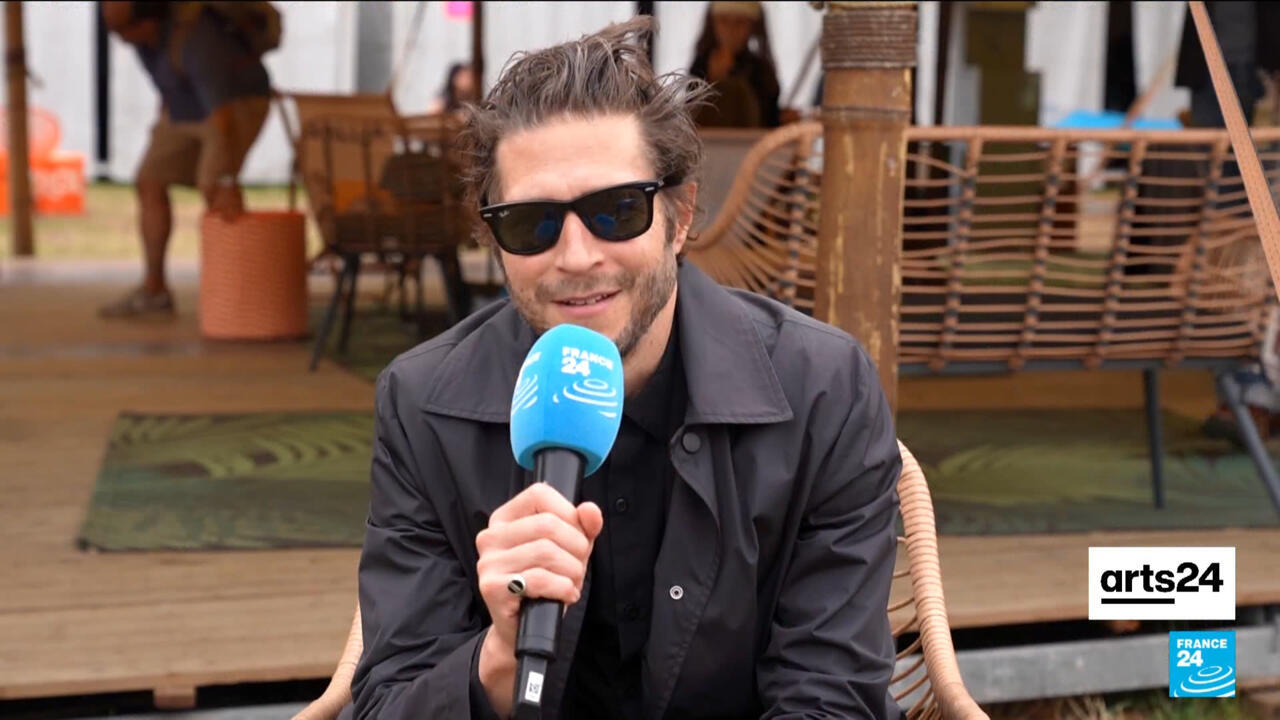 A man in sunglasses holds a France 24 microphone while seated outdoors.