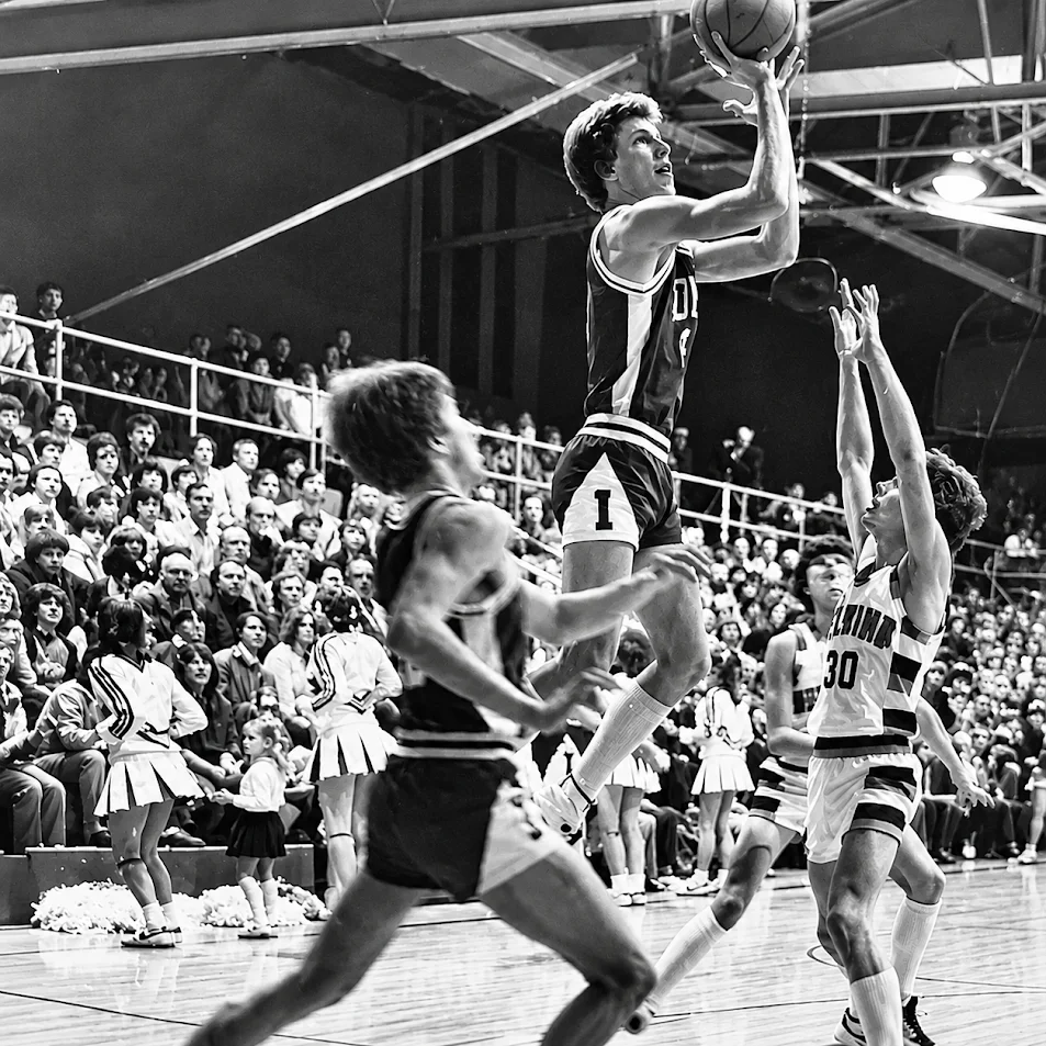 Black-and-white basketball player jumps toward the hoop as defenders contest the shot before a packed crowd.