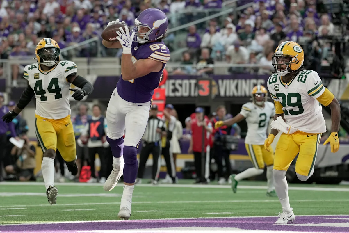 Football player in purple catches a pass as two defenders in green and yellow pursue.