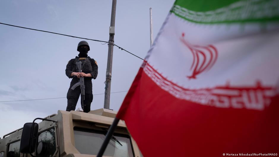 Armed soldier stands atop an armored vehicle behind a large Iranian flag.