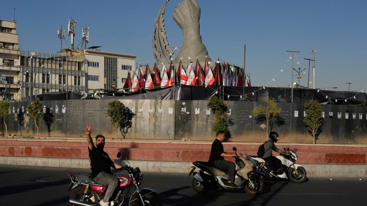 Motorcyclists pass Iranian flags and a large monument along a city street.
