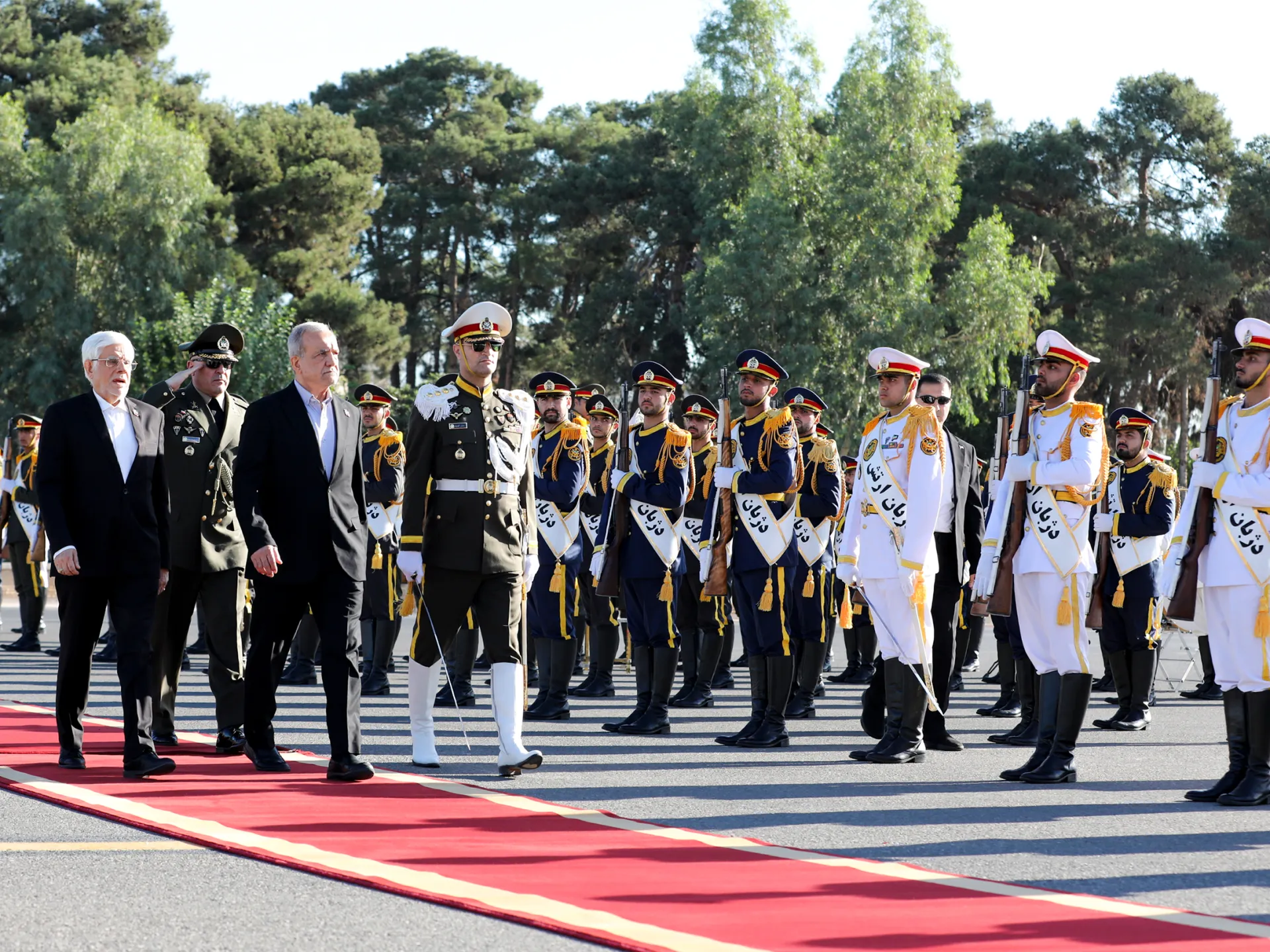 Officials walk past uniformed ceremonial guards standing at attention along a red carpet outdoors.
