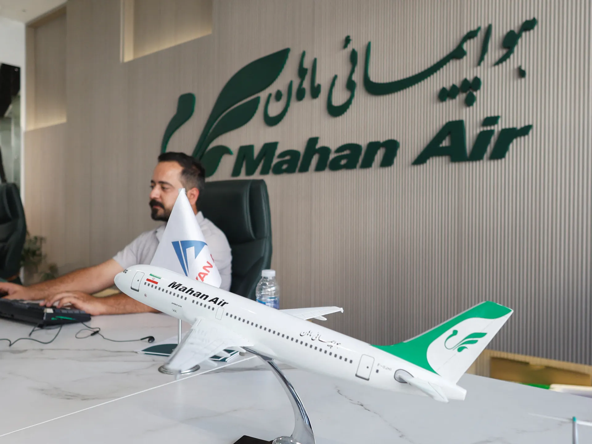 A man sits at a desk beside a large model passenger aircraft in an office.