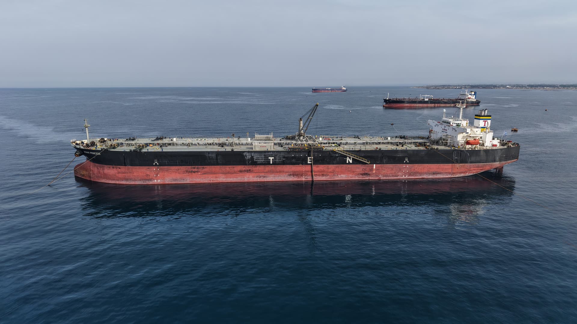 A large tanker sits at anchor on calm blue water, with other ships visible in the distance.
