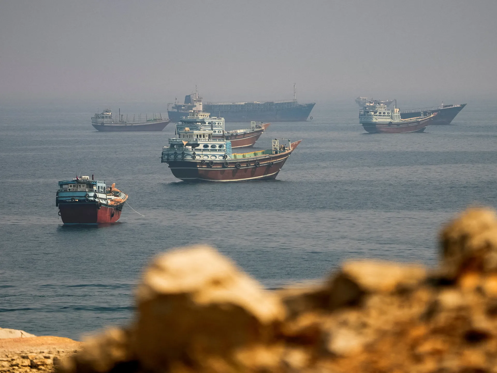 Several traditional cargo boats float on hazy water near a rocky shoreline.