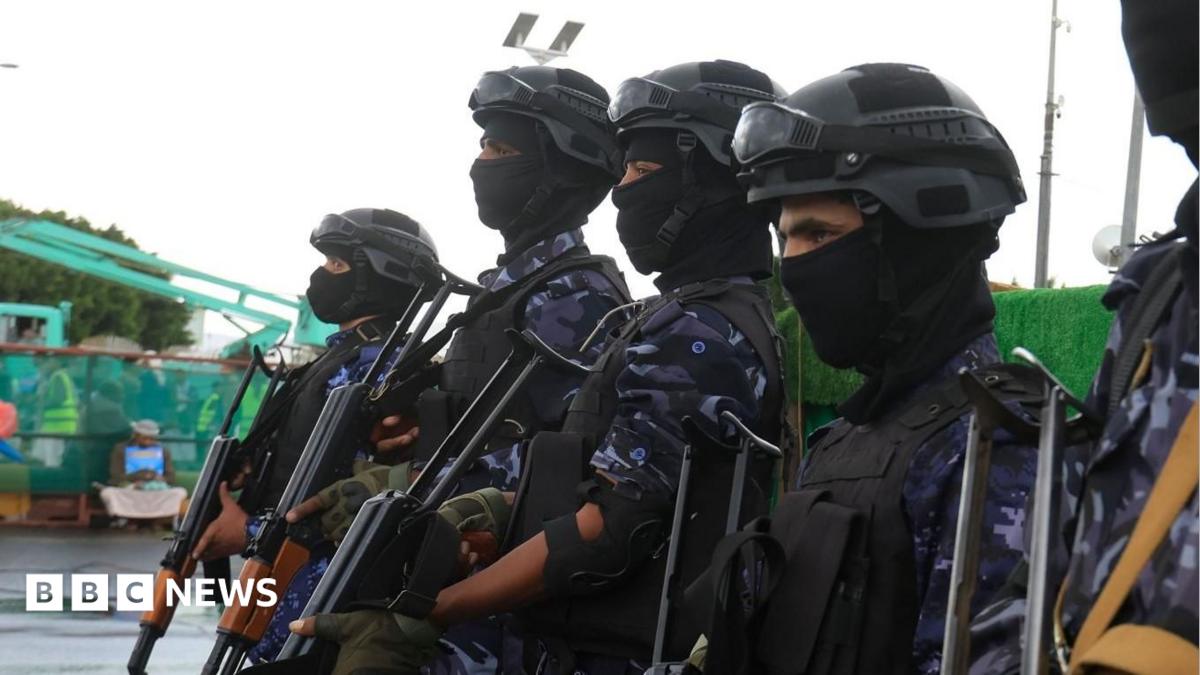 Four masked armed personnel in helmets and camouflage uniforms stand holding rifles outdoors.