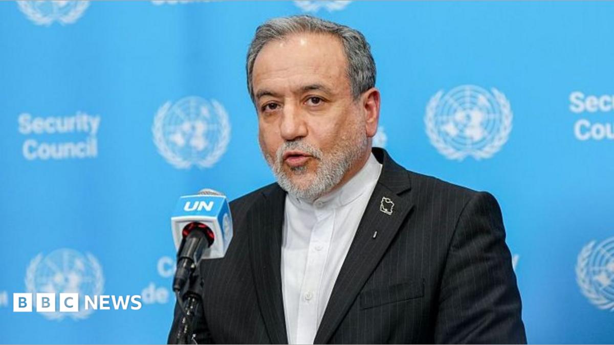 A gray-haired man in a dark suit speaks into a UN microphone against a blue Security Council backdrop.