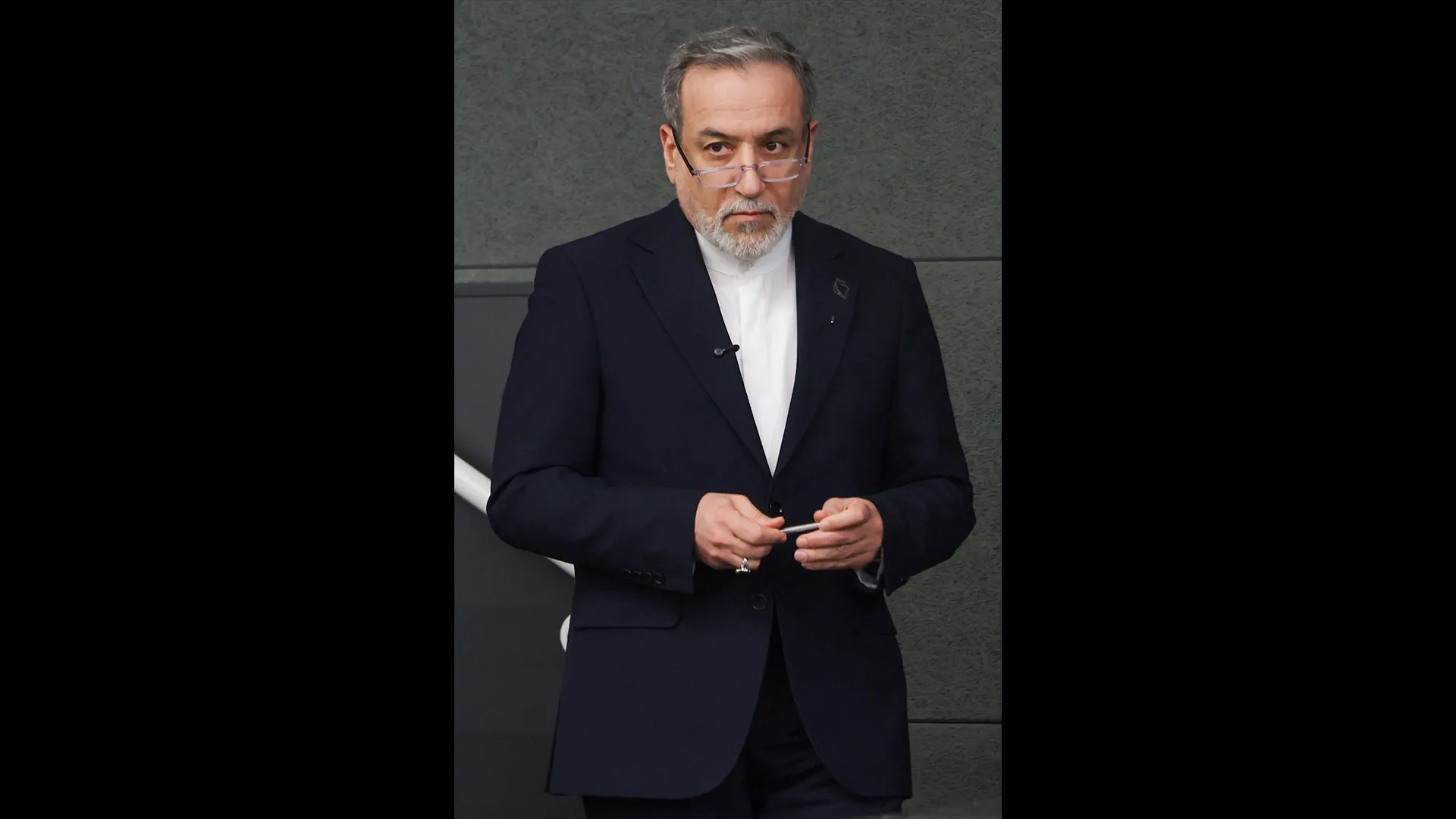 A gray-haired, bearded man in a dark suit and glasses holds a pen against a gray wall.