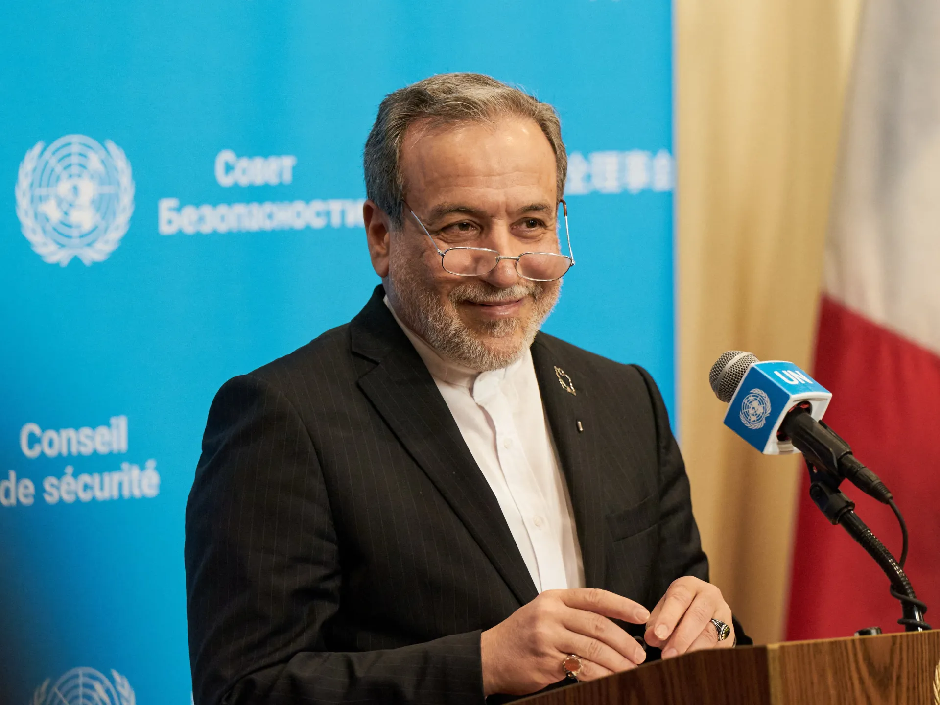 A man in a dark suit speaks at a podium beside a UN microphone, with flags behind him.