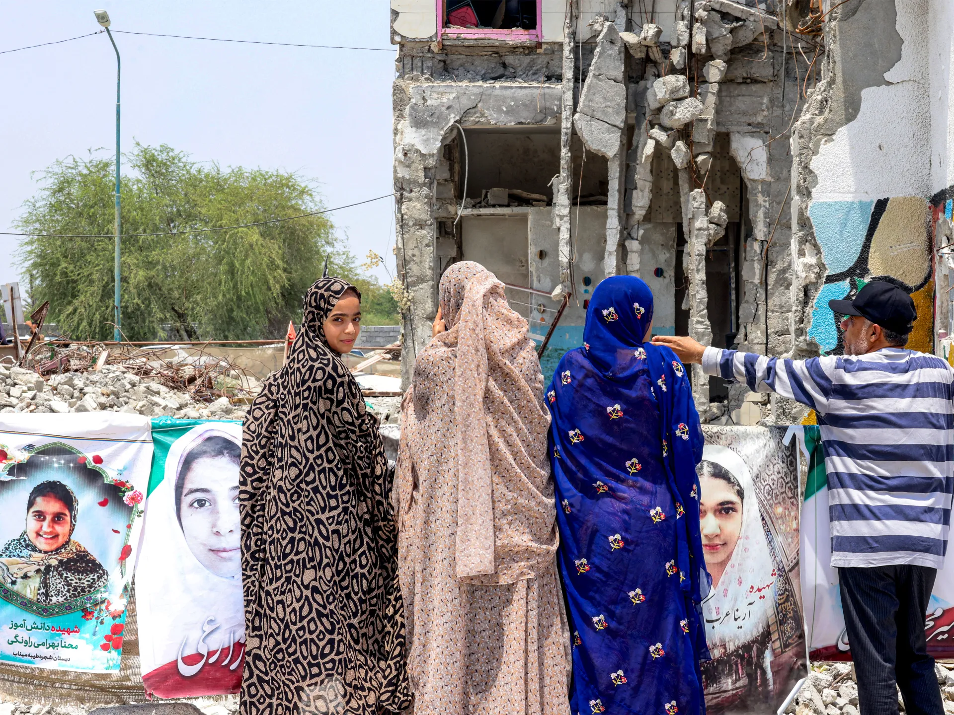 Three women and a man stand beside portraits in front of a heavily damaged building.