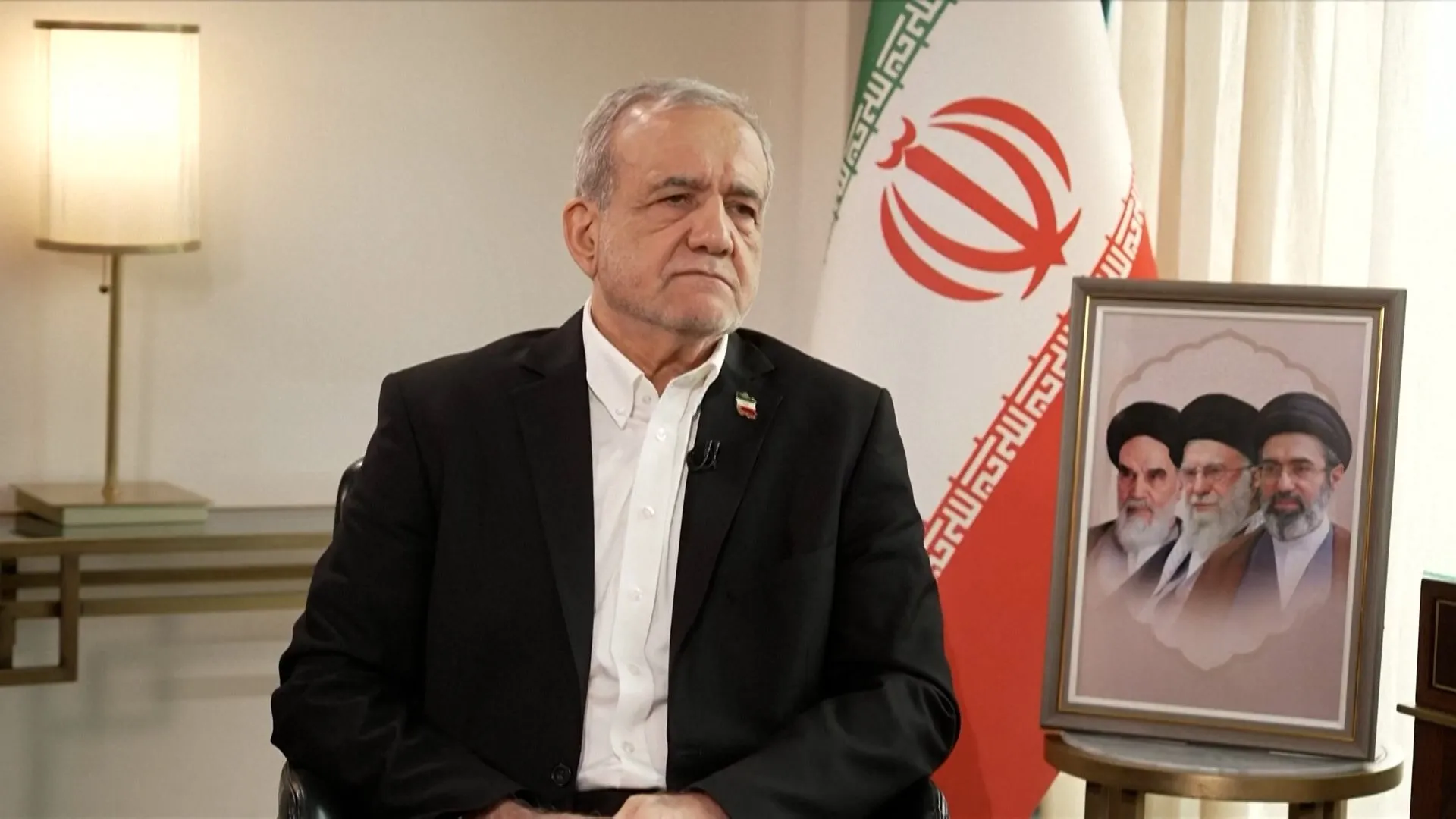 Man in a dark suit seated beside an Iranian flag and framed clerical portrait