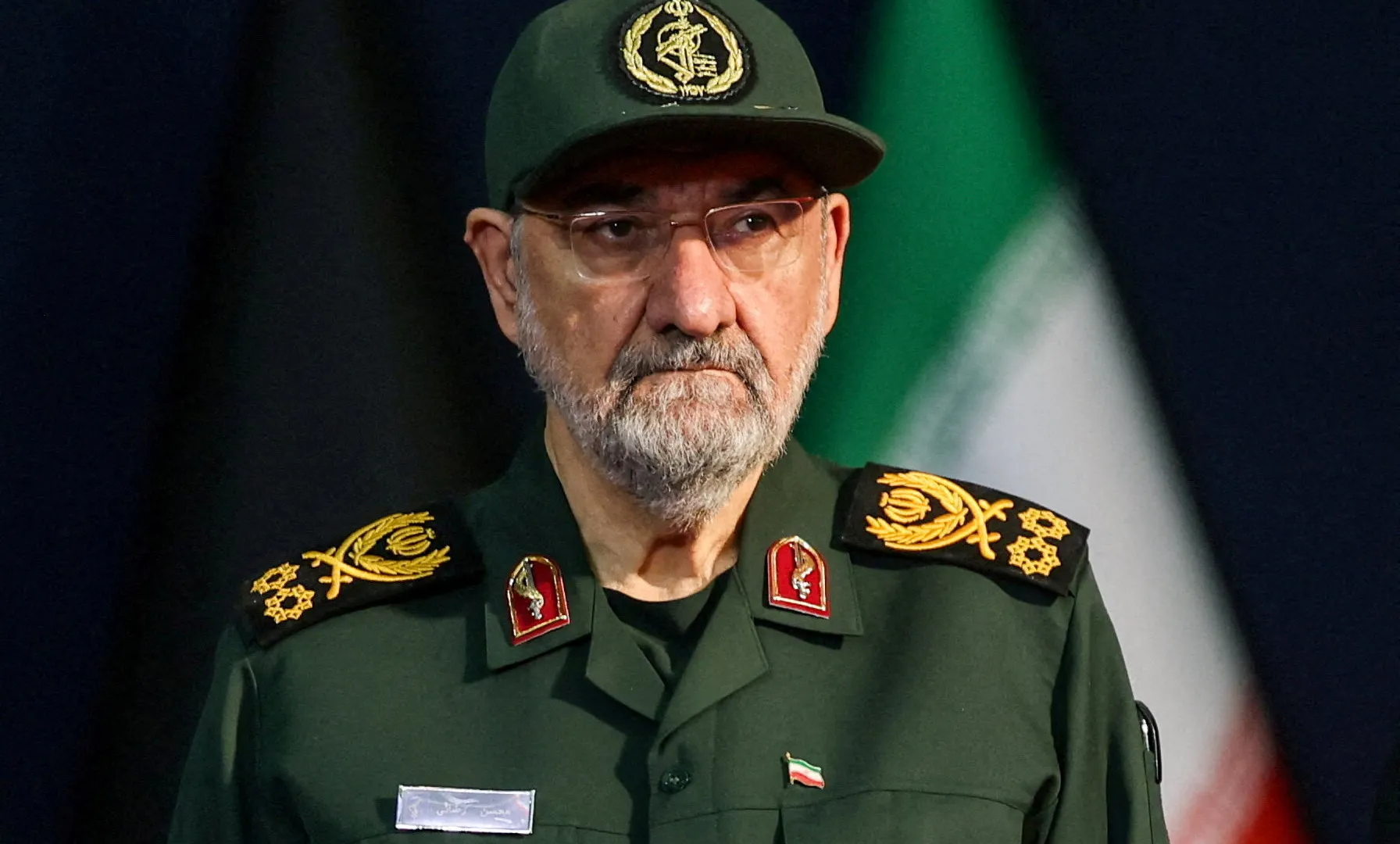Bearded Iranian military official in green uniform and cap pictured before an Iranian flag.