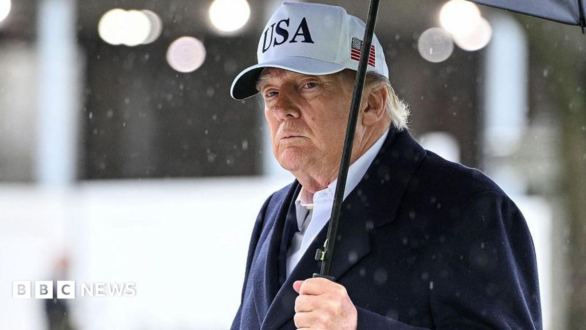 Donald Trump wears a white USA cap and dark coat, holding an umbrella in the rain.