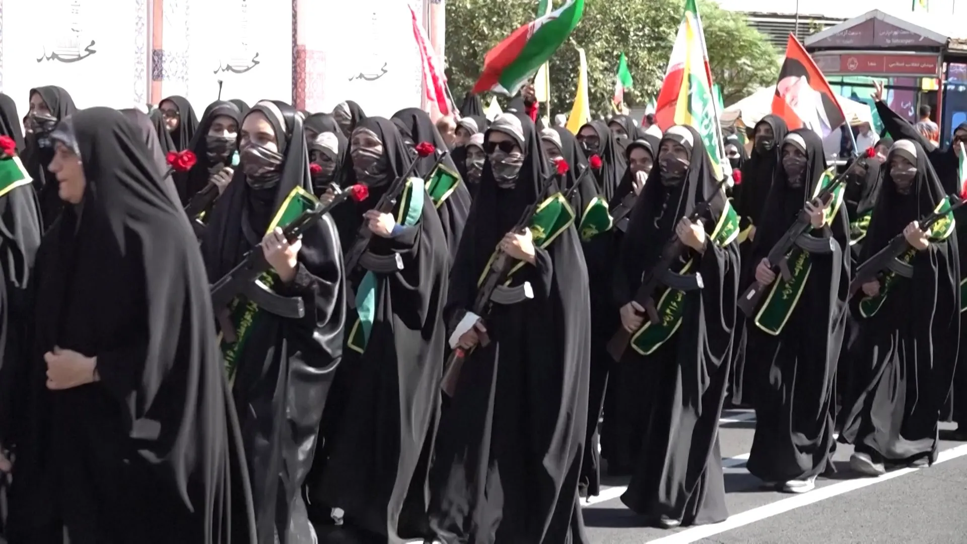 Women in black veils march carrying rifles, roses, and green flags in a street procession.