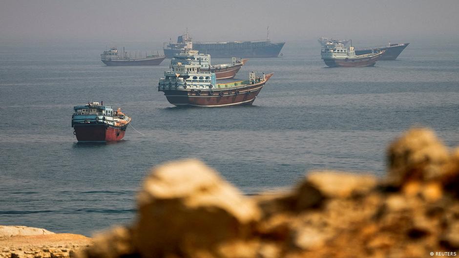 Several fishing boats sit offshore beyond a rocky shoreline under hazy skies.