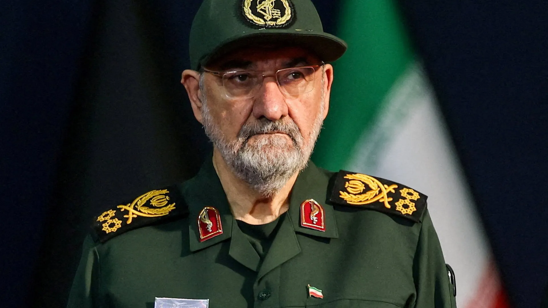 Bearded military officer in green uniform and cap posed before an Iranian flag.