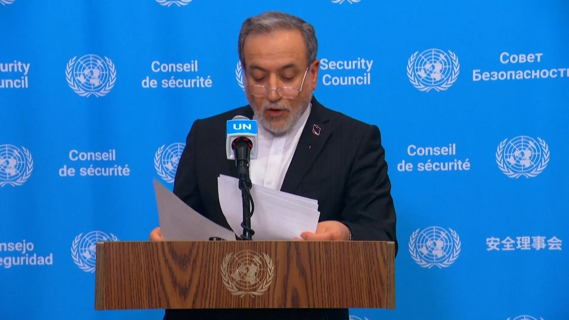 A gray-haired man reads from papers at a microphone behind a wooden United Nations podium.