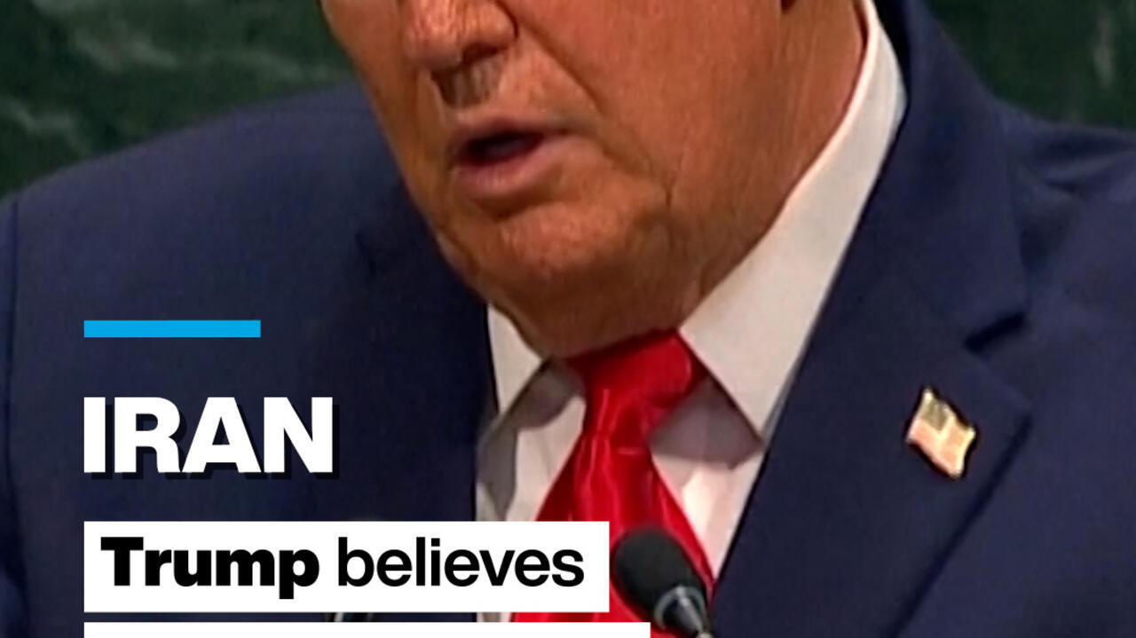 Close-up of a suited man wearing a red tie and U.S. flag lapel pin speaking at a microphone