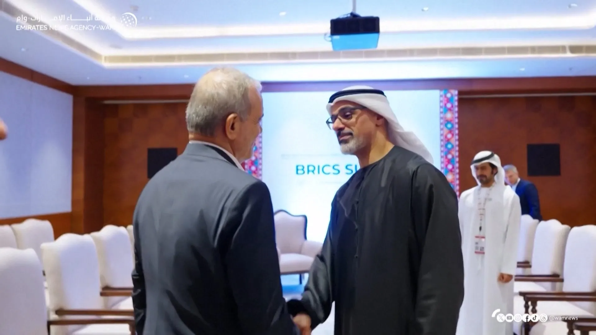 Two men shake hands during a BRICS summit event, with attendees and conference seating behind them.