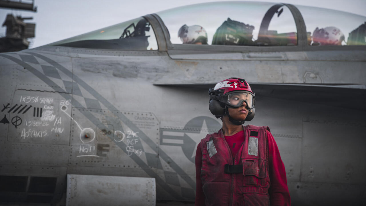 Flight deck crew member in red gear stands beside a weathered military jet.