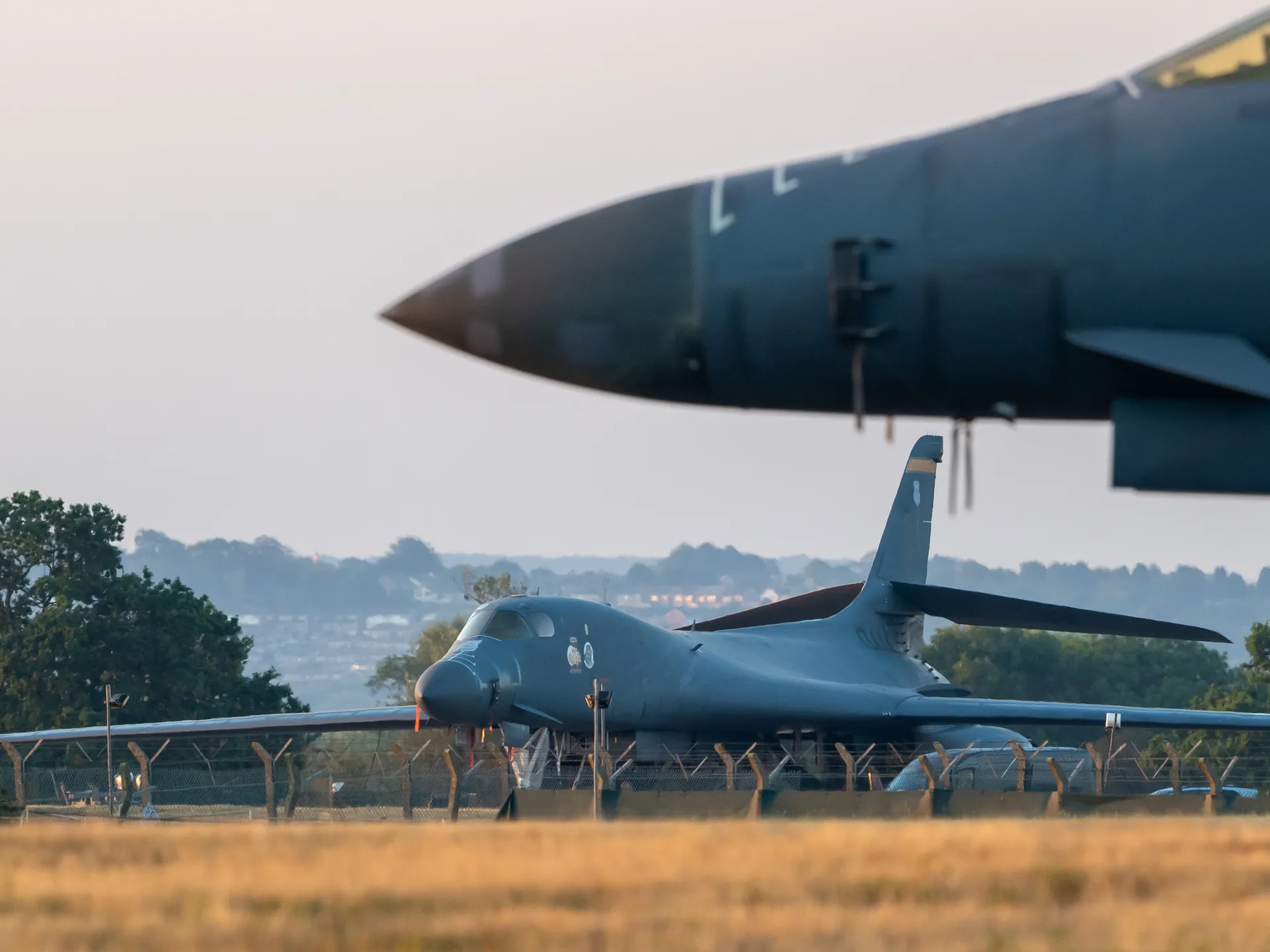 Military aircraft parked behind security fencing with another aircraft passing overhead at an airfield.