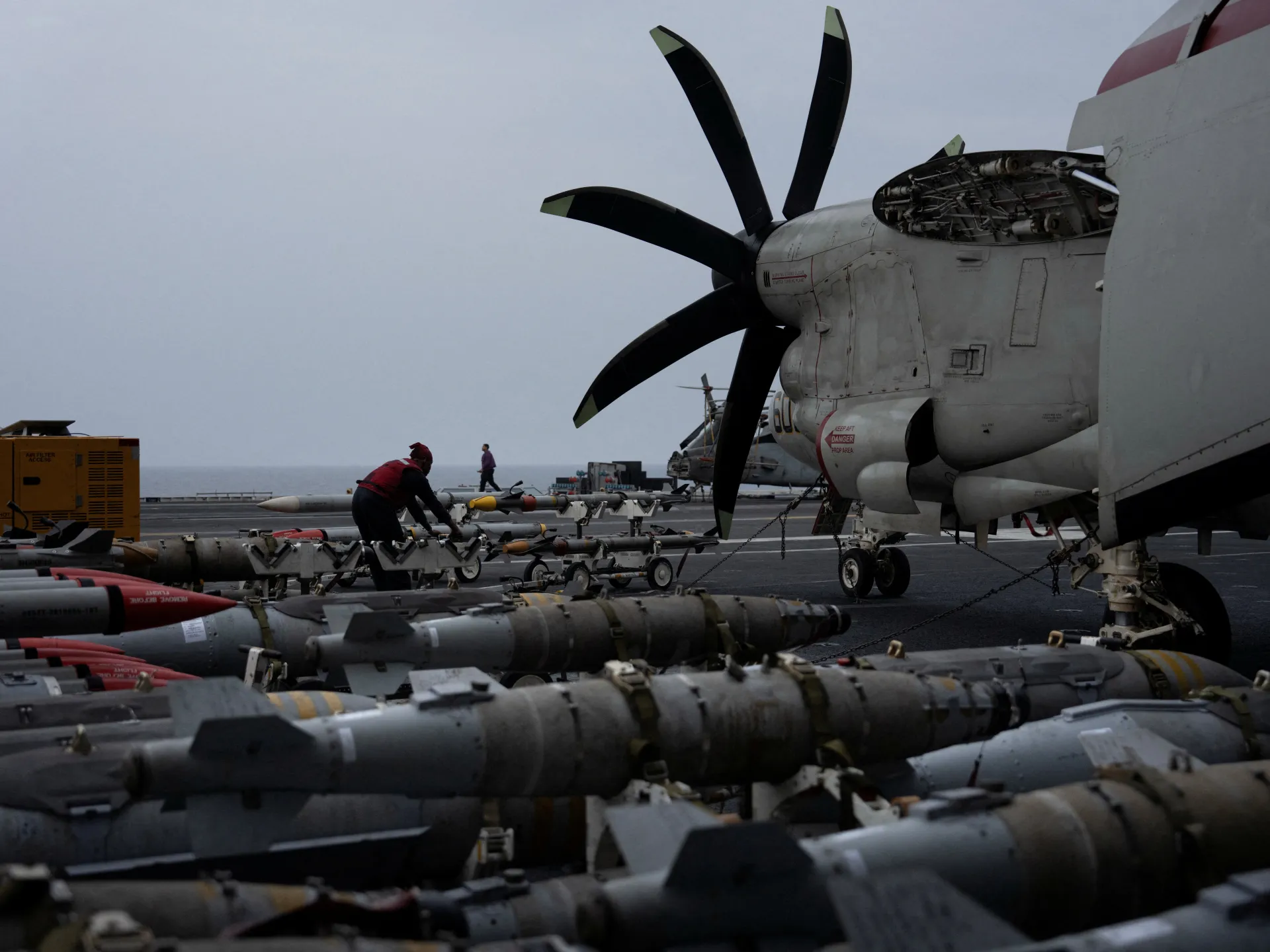 Military aircraft with exposed propeller sits beside rows of missiles on a ship deck.