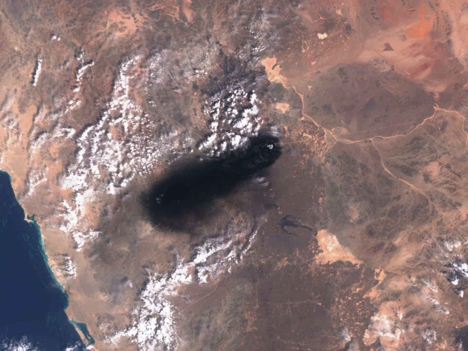 Satellite view of arid terrain, rugged snow-capped mountains, coastline, and a large dark cloud shadow.