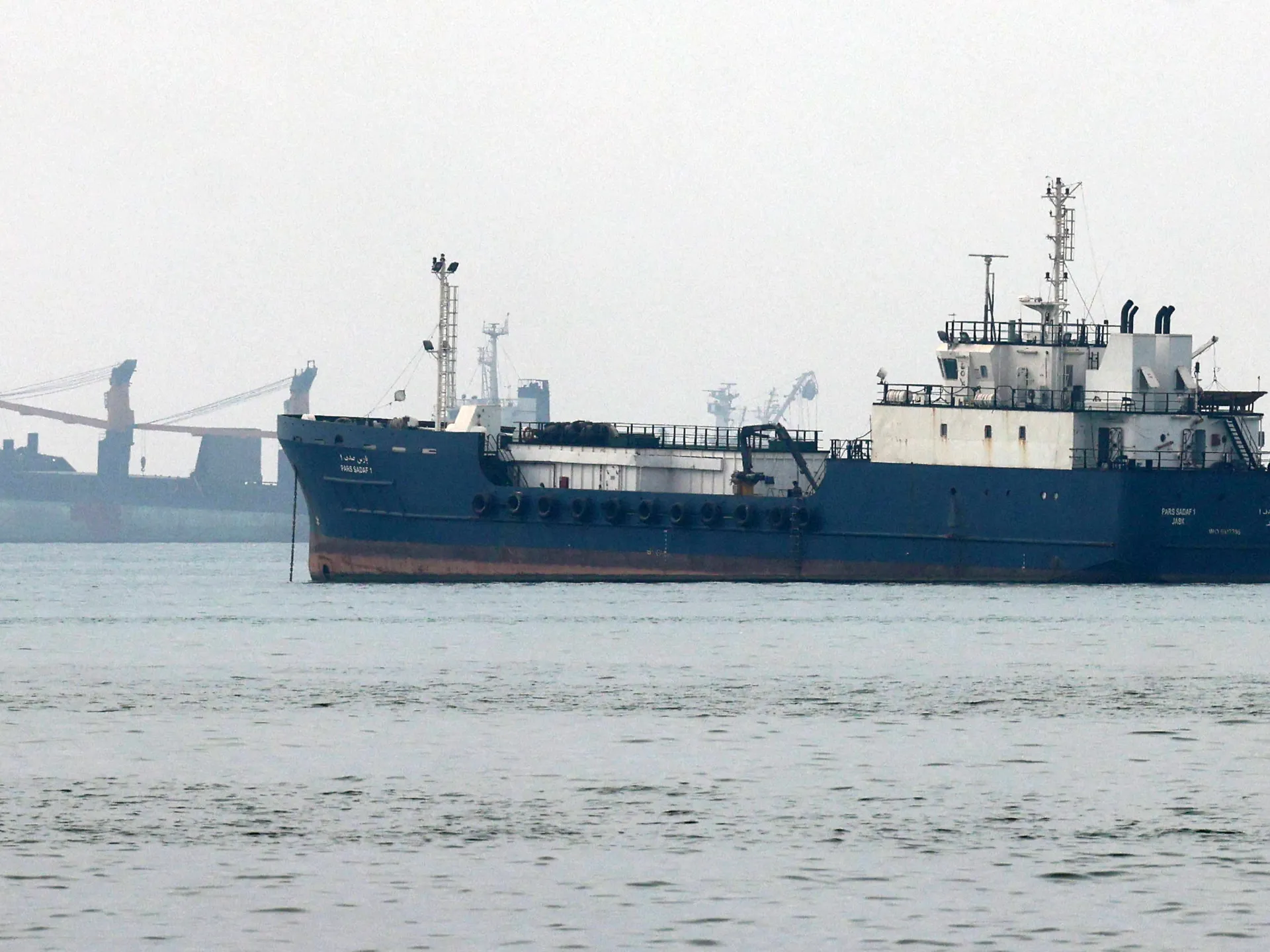 Large blue cargo ship anchored near port cranes under a hazy sky