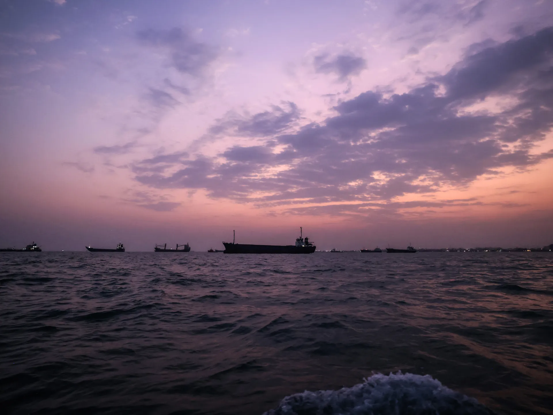Cargo ships silhouetted on dark water beneath a pink and purple dusk sky.