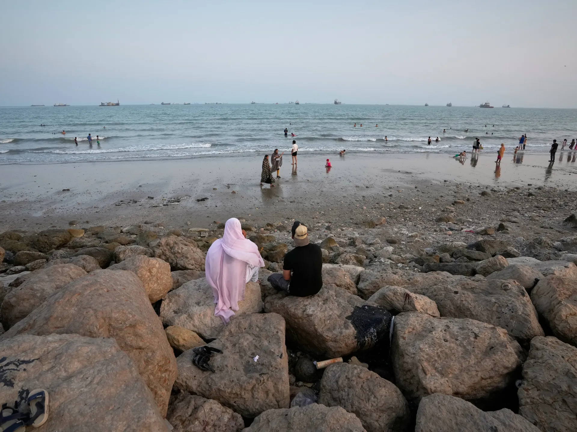 People sit on rocks overlooking swimmers, waves, and numerous ships on a hazy sea horizon.