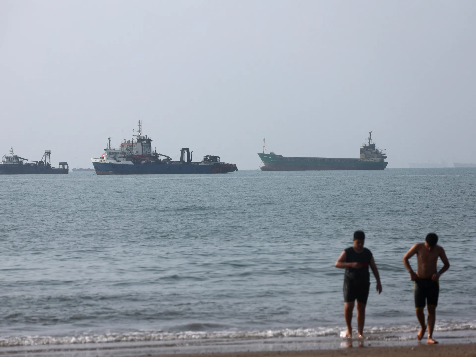 Cargo and fishing vessels offshore while two men walk through shallow water at a beach.