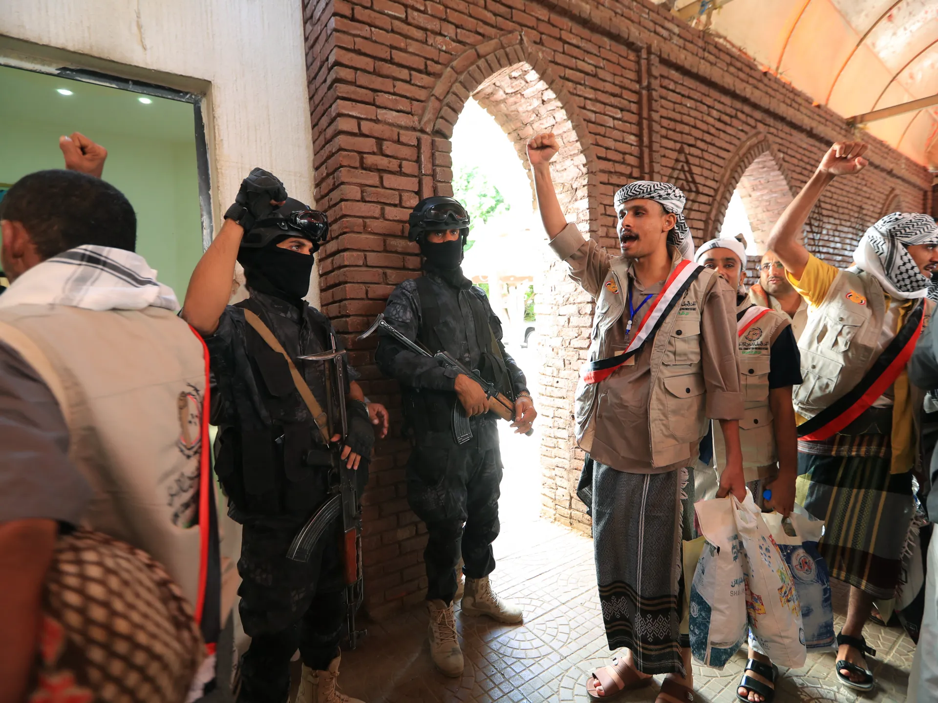 Masked armed men stand beside civilians raising fists inside a brick-arched building.