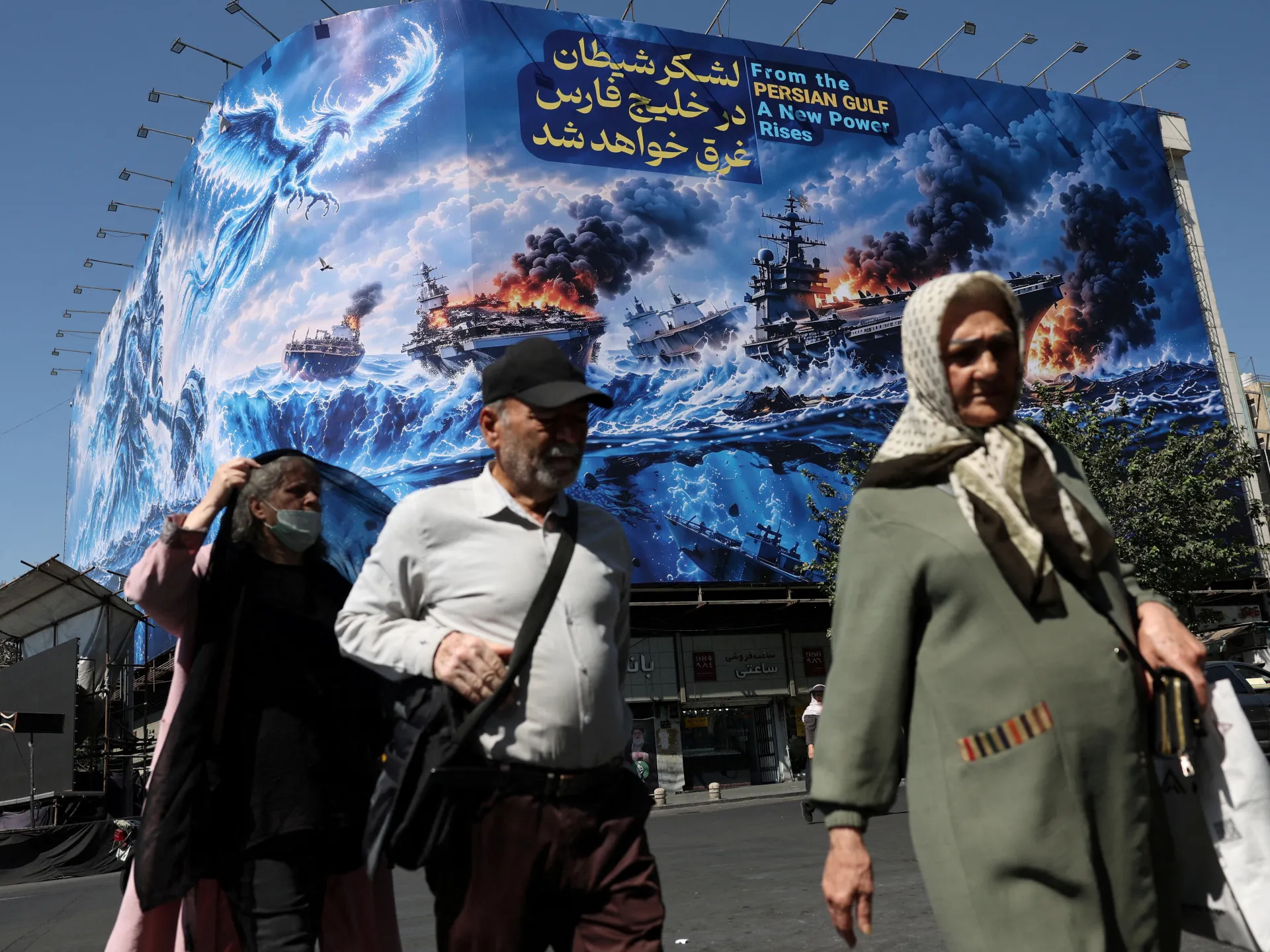 Pedestrians pass a billboard depicting burning warships beneath Persian and English text about the Persian Gulf.