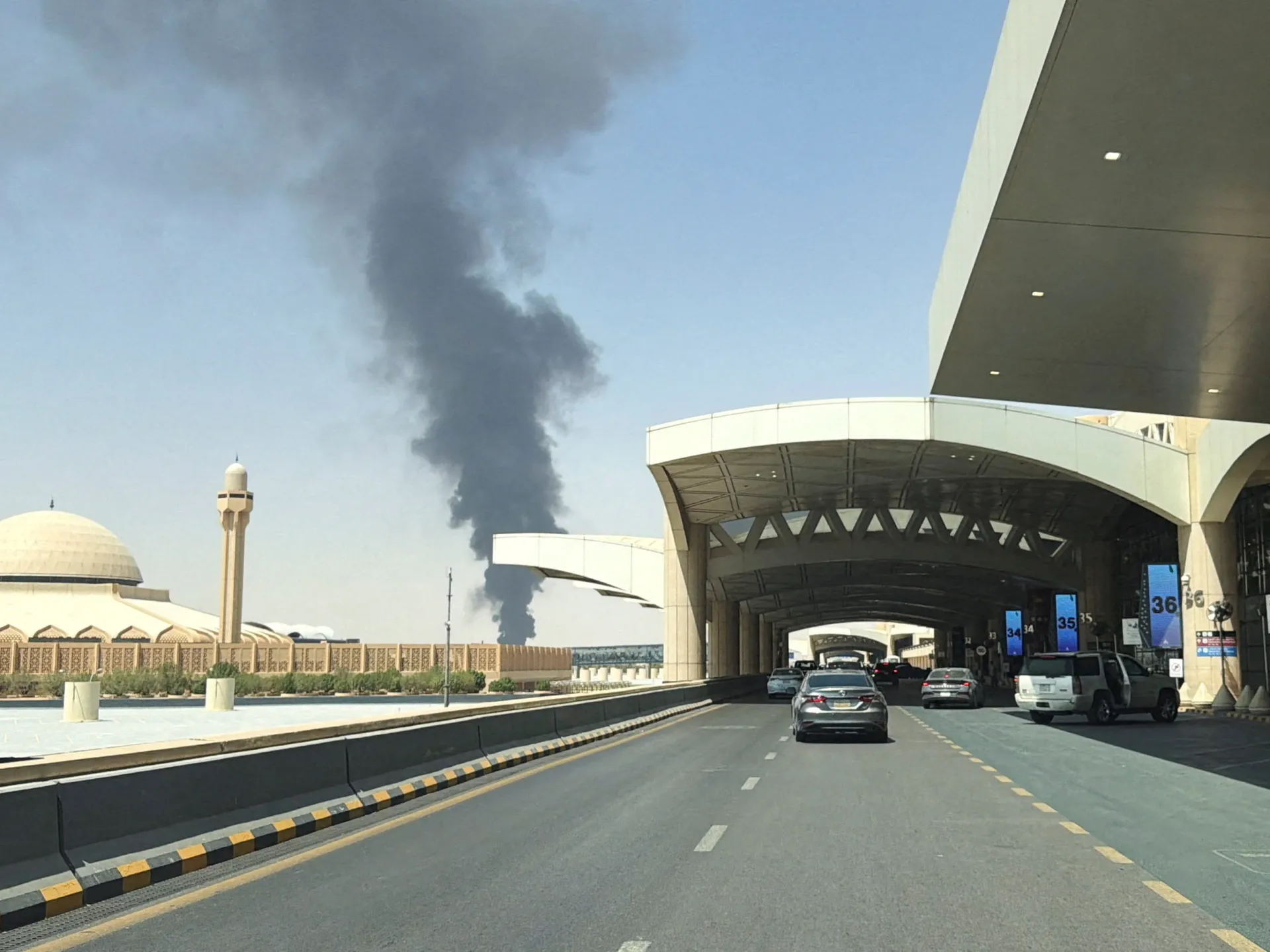 Smoke plume rises behind a mosque beside a busy airport terminal roadway.