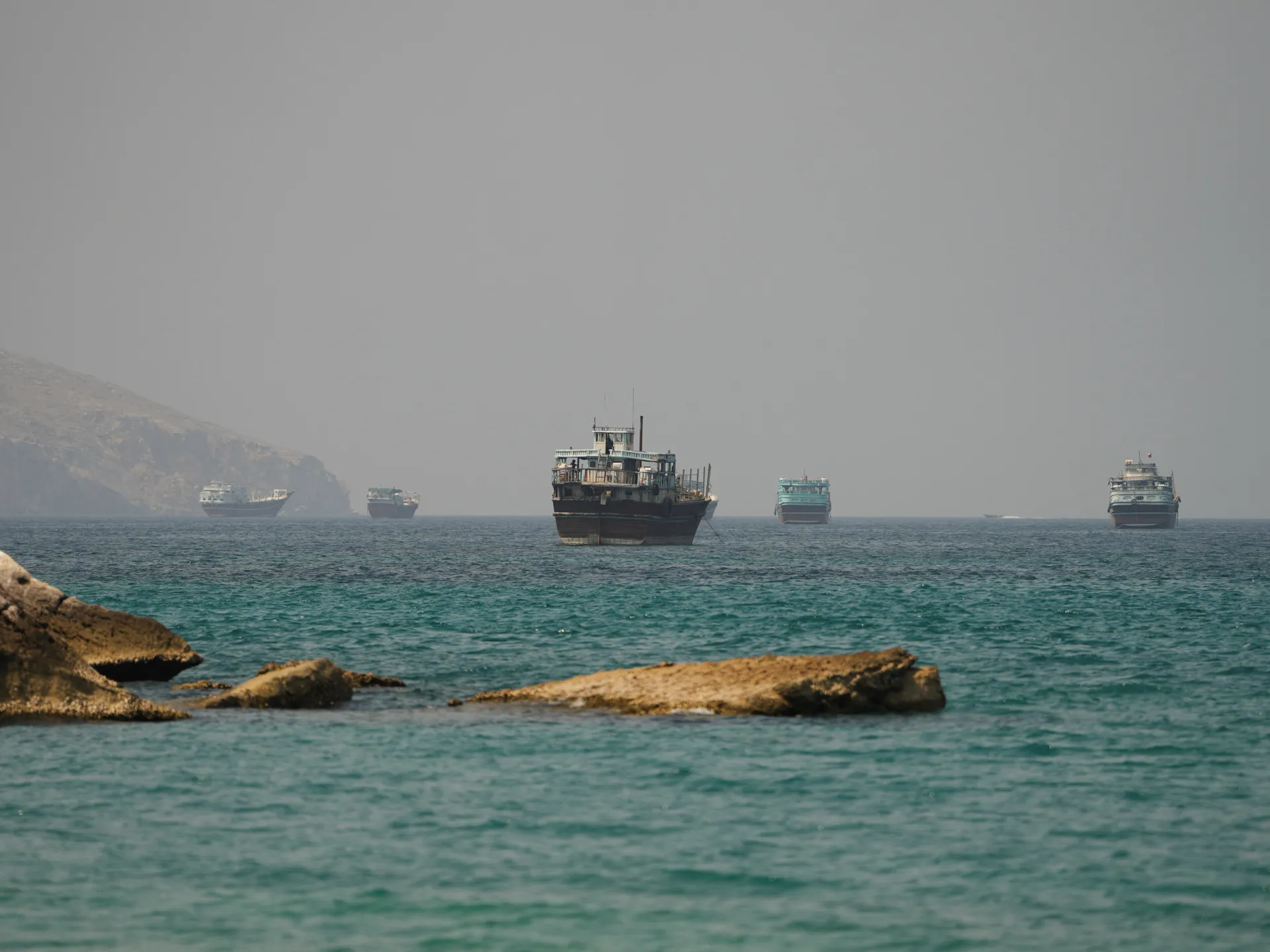 Several traditional boats float offshore beside rocky outcrops beneath a hazy sky.
