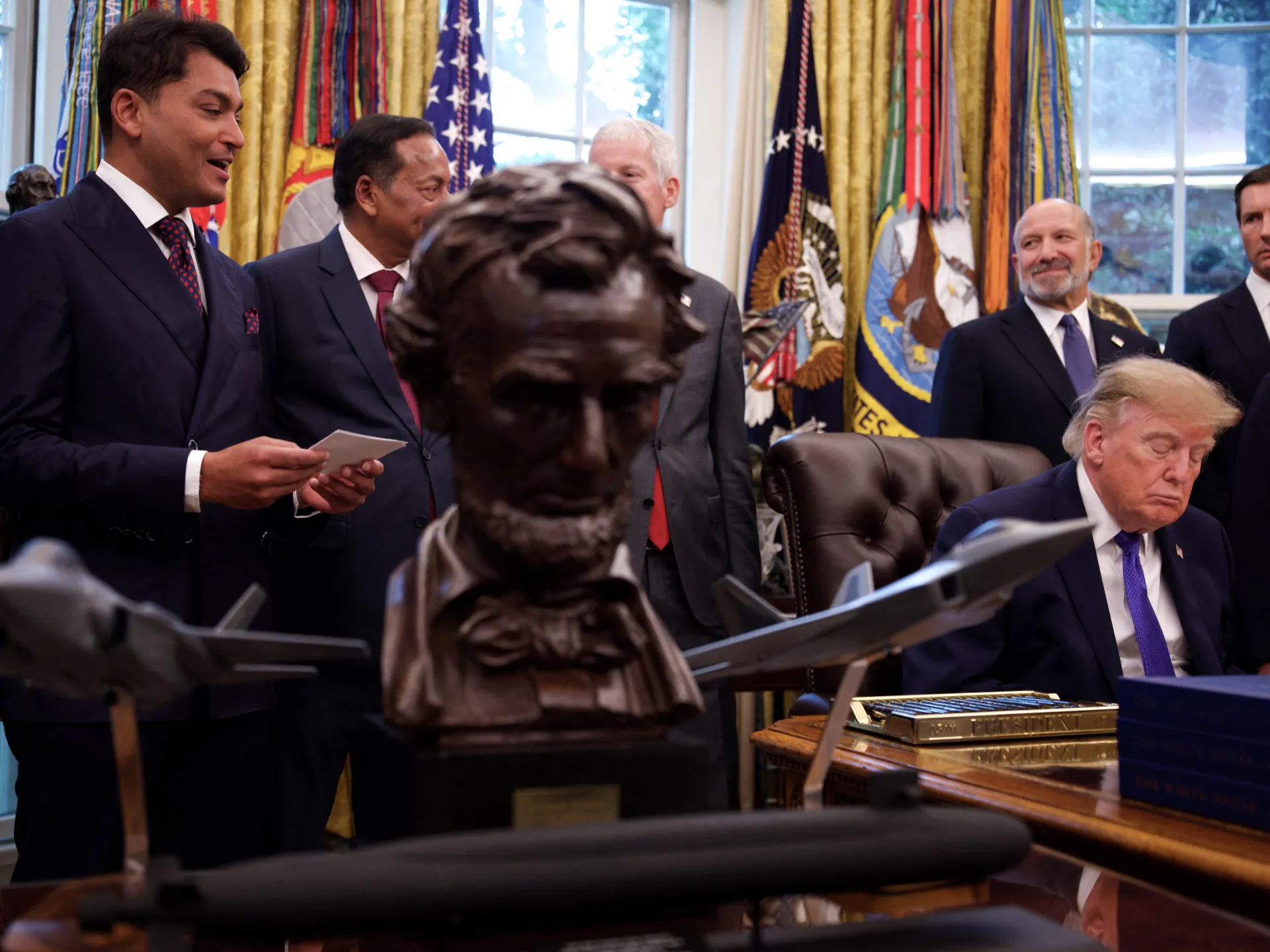 A seated man looks down at a desk as officials stand behind him and a bust fills the foreground.
