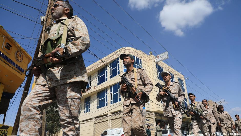 Armed men in desert camouflage walk through a city street beneath overhead power lines.