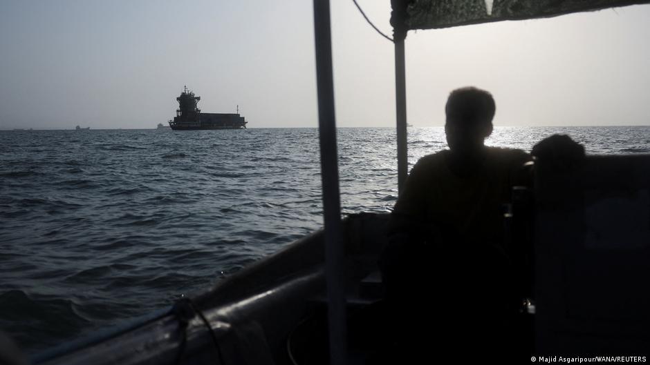 Silhouetted passengers aboard a small boat watch a large vessel offshore under a hazy sky.