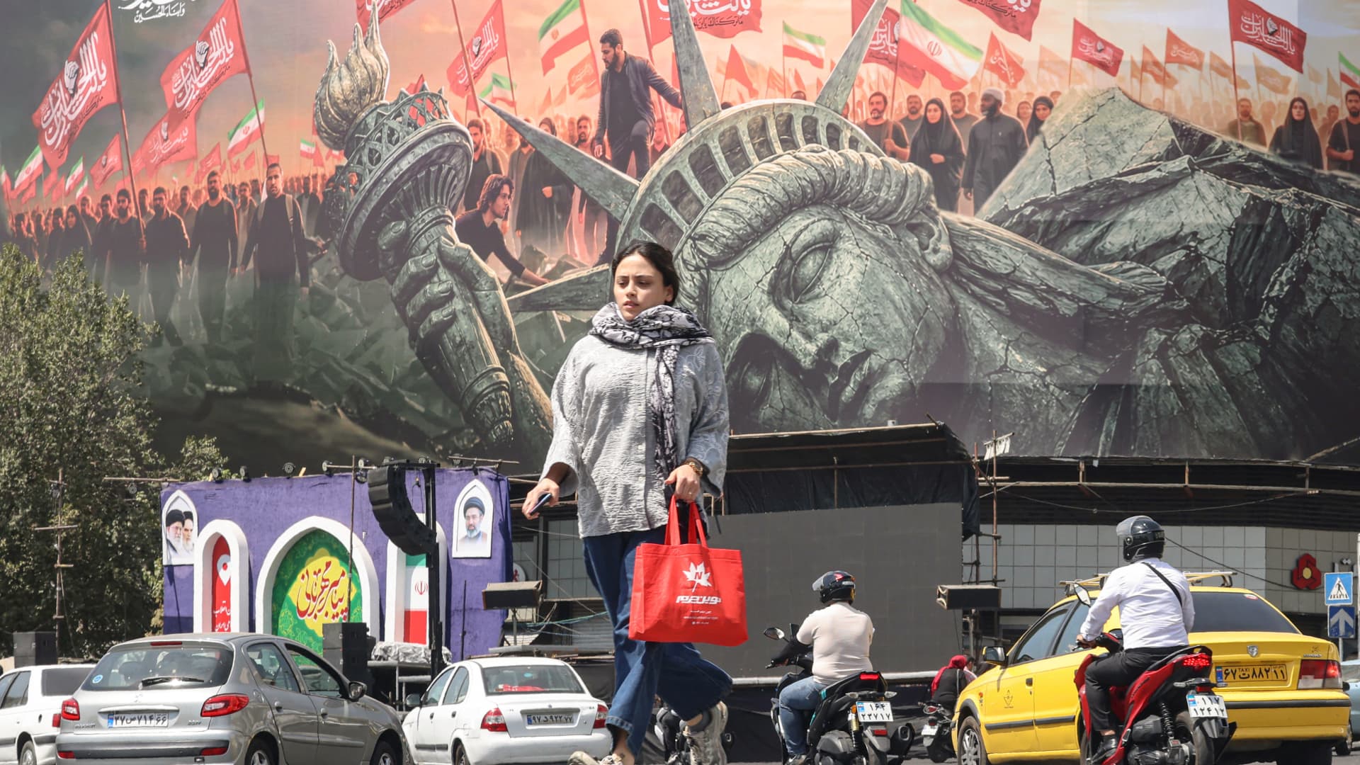 Woman carrying a red shopping bag walks past traffic and a large political mural with flags.