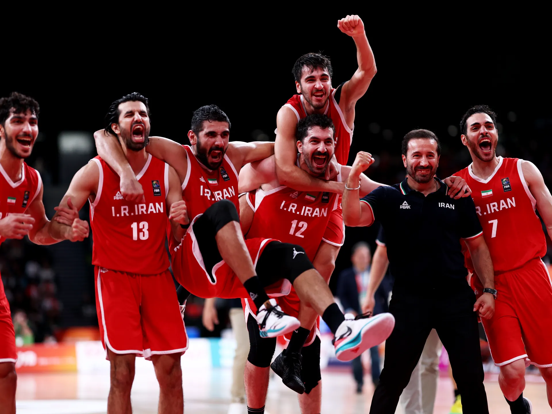 Iranian basketball players in red uniforms celebrate together, with one player lifted and raising his fist.