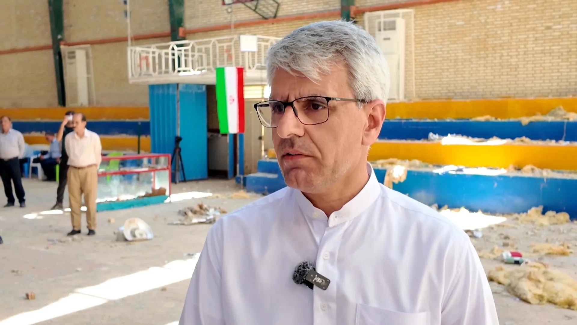 Gray-haired man in glasses speaking near an Iranian flag inside a damaged indoor venue