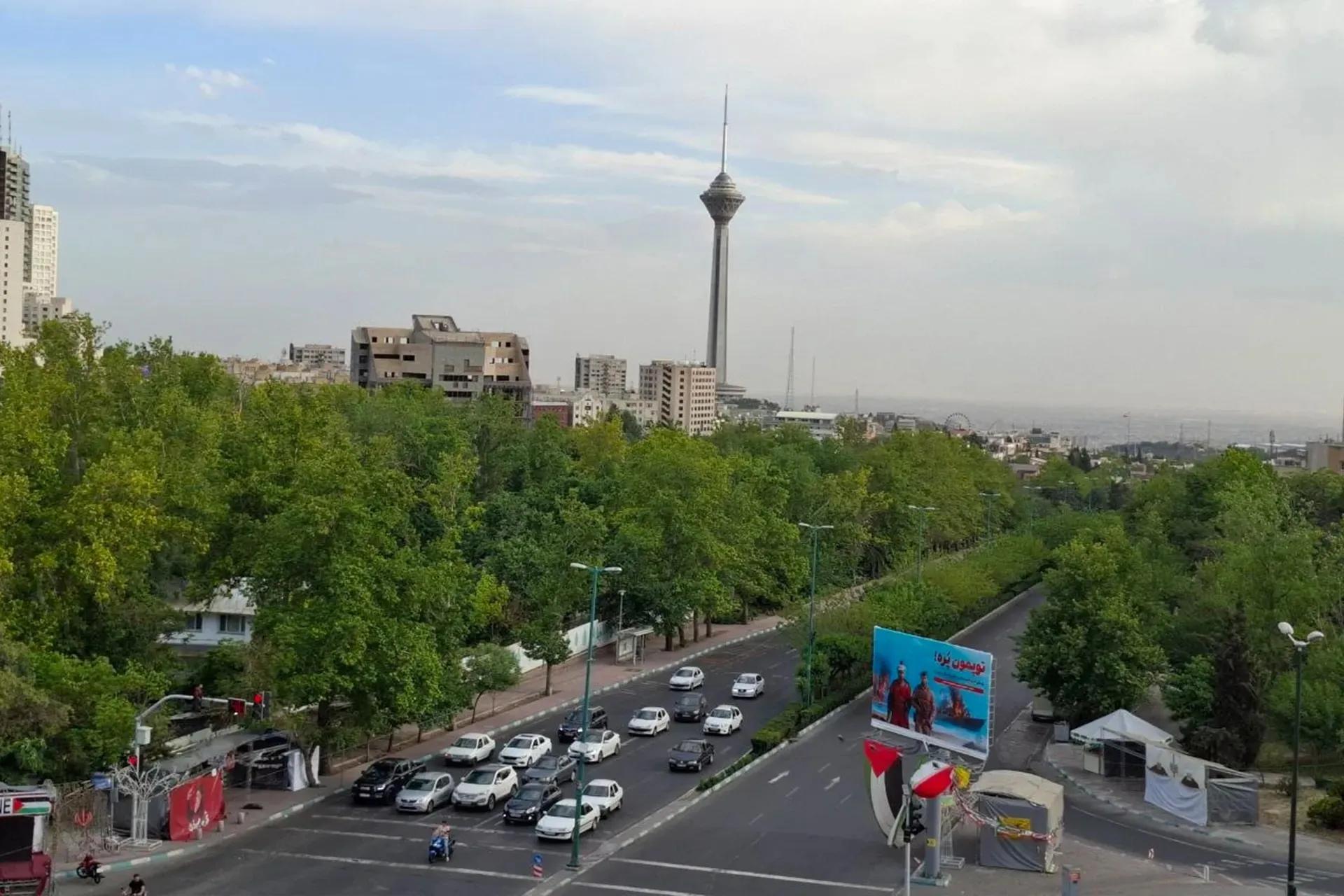 A tree-lined city road with cars in traffic, a billboard, and a tall communications tower under a hazy sky.