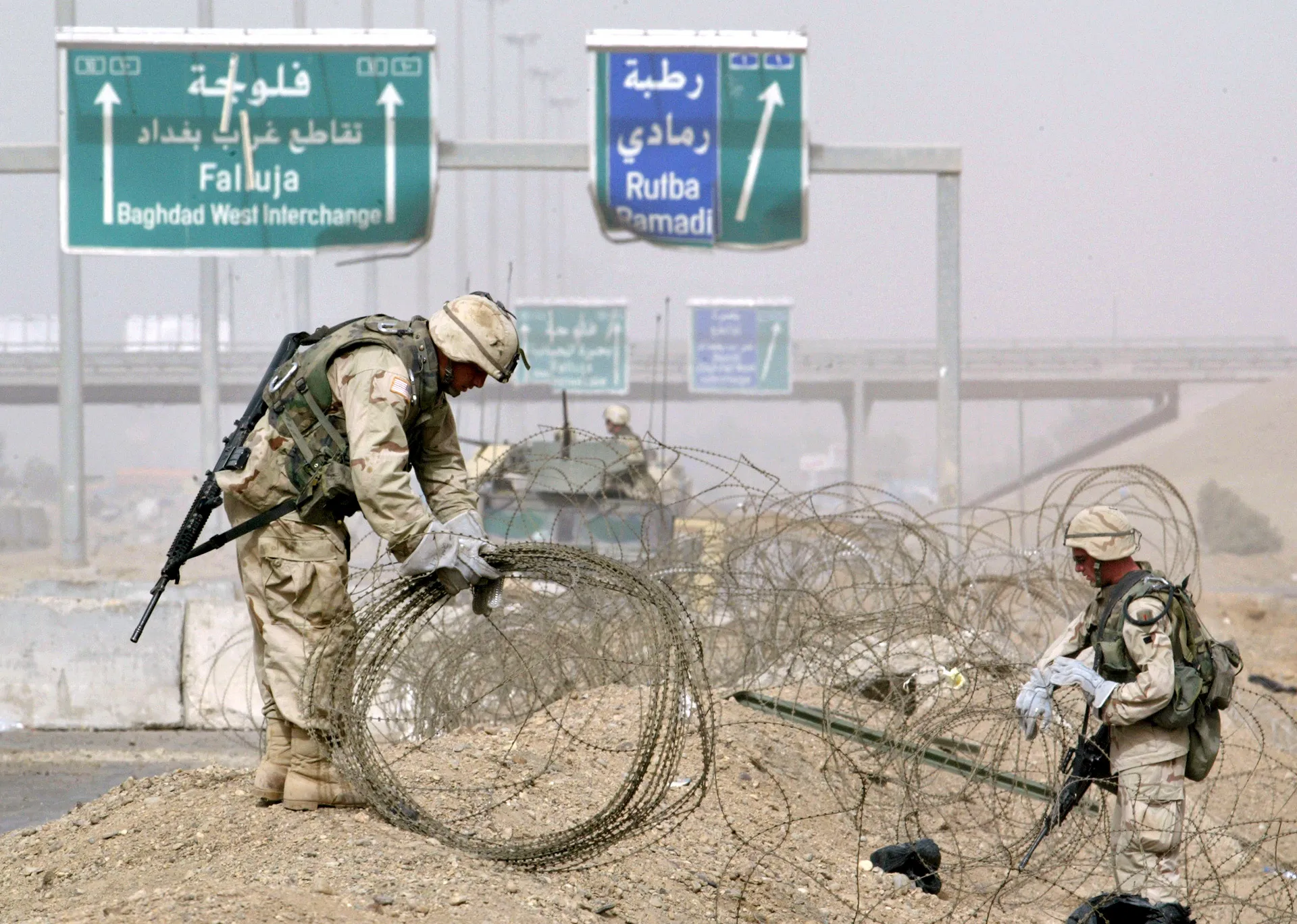 Soldiers in desert camouflage handle concertina wire beneath multilingual highway signs.